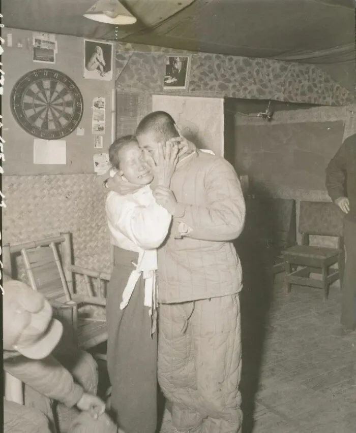 Two people embracing with joy in a historical photograph, with a dartboard and pictures on the wall behind them.