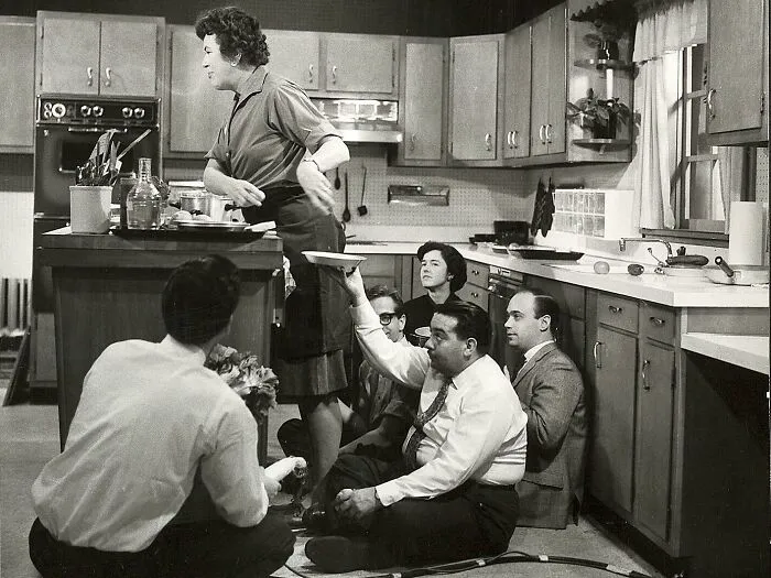Historical photo: a woman in a kitchen being filmed, surrounded by a film crew sitting on the floor.