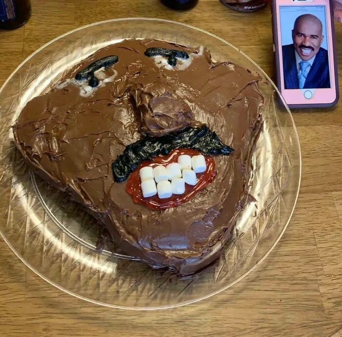 Funny cake fail with a chocolate-frosted face and marshmallow teeth, next to a phone displaying a reference image.