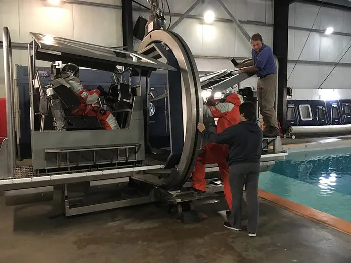 Three people in safety gear operating a large metal training device near an indoor pool for online group safety facts.