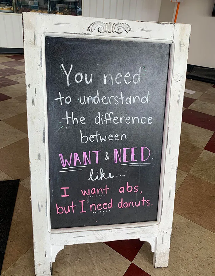 Funny sign about the difference between want and need, stating, "I want abs, but I need donuts," written on a chalkboard.