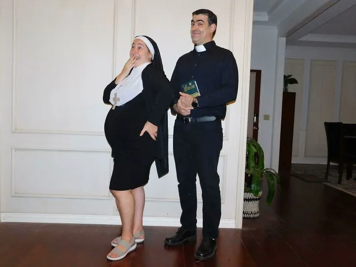 Couple with simple Halloween costumes dressed as a pregnant nun and a priest holding a Bible indoors.