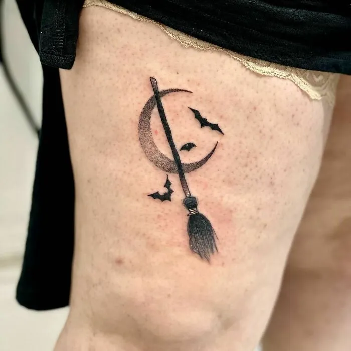 Spooky Halloween tattoo featuring a crescent moon, broomstick, and bats on the thigh.