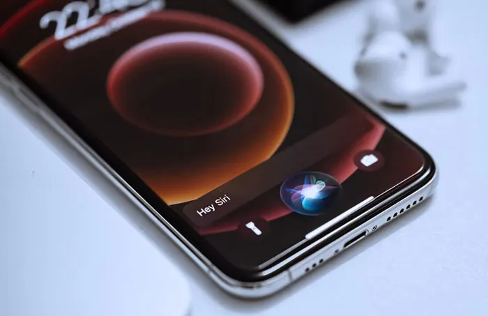 A smartphone with a dark-themed screen displaying "Hey Siri," placed on a white surface with earbuds nearby.