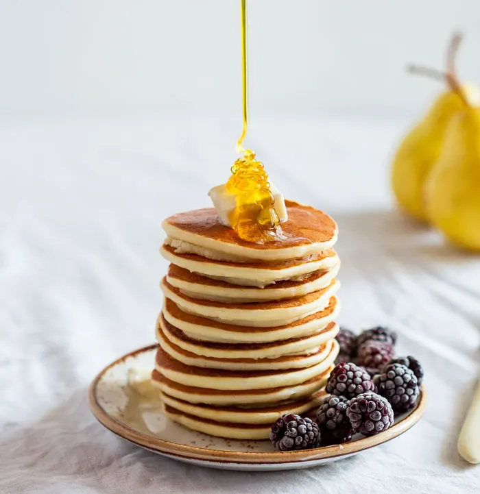 Stack of pancakes with syrup, butter, and blackberries, on a plate with pears in the background, related to dark jokes.