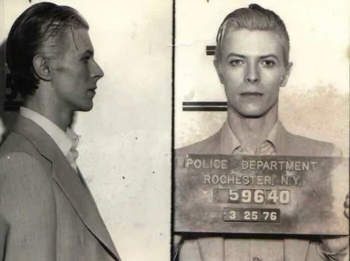 A historical photo showing a mugshot from 1976, featuring a person in a suit with slicked-back hair.