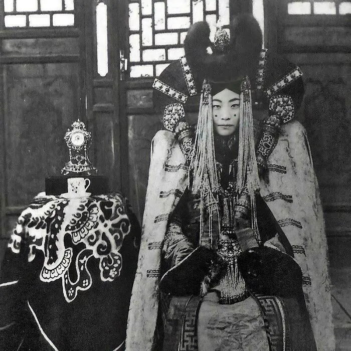 Historical photo of a person in traditional ornate attire, seated beside an intricately decorated table with a clock.