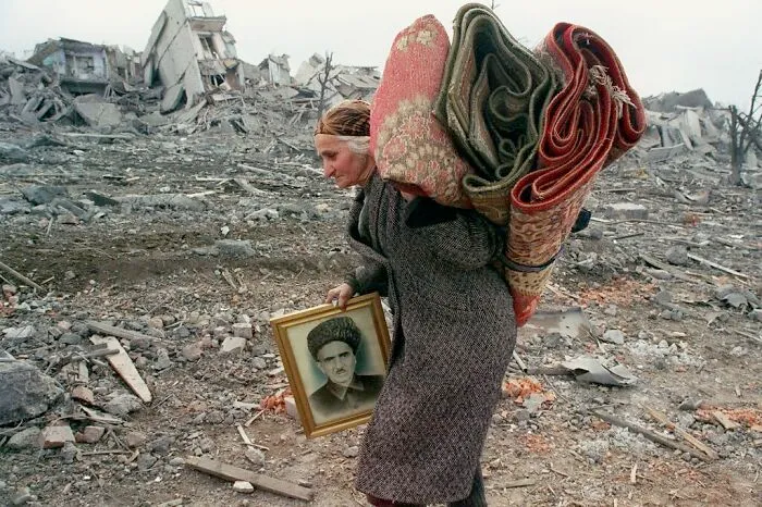 Woman carrying rugs and a framed photo through rubble, highlighting important historical perspectives.