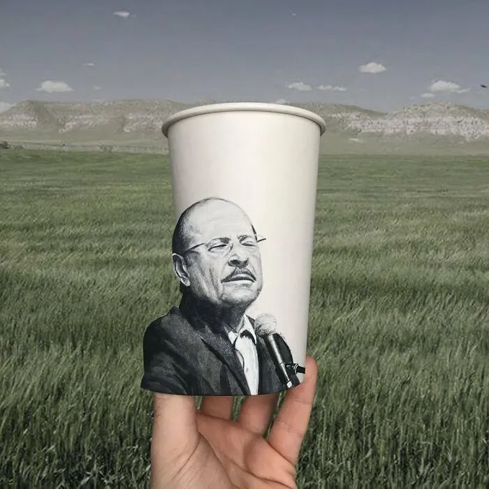Hand holding a paper cup with a detailed illustration of a man speaking into a microphone outdoors in a grassy field.