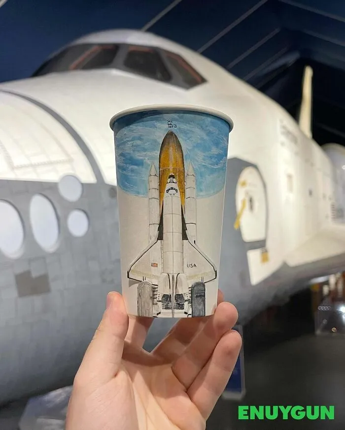 Hand holding paper cup with detailed space shuttle illustration in front of a real space shuttle, showcasing travel-themed art.