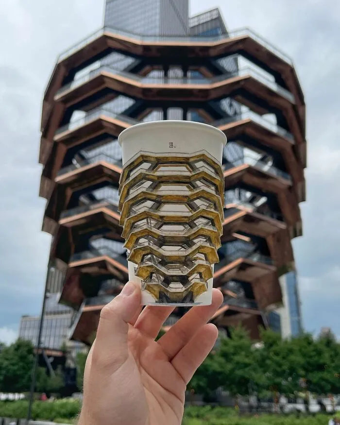 Hand holding a paper cup with detailed illustrations matching a large architectural structure outdoors during daytime.