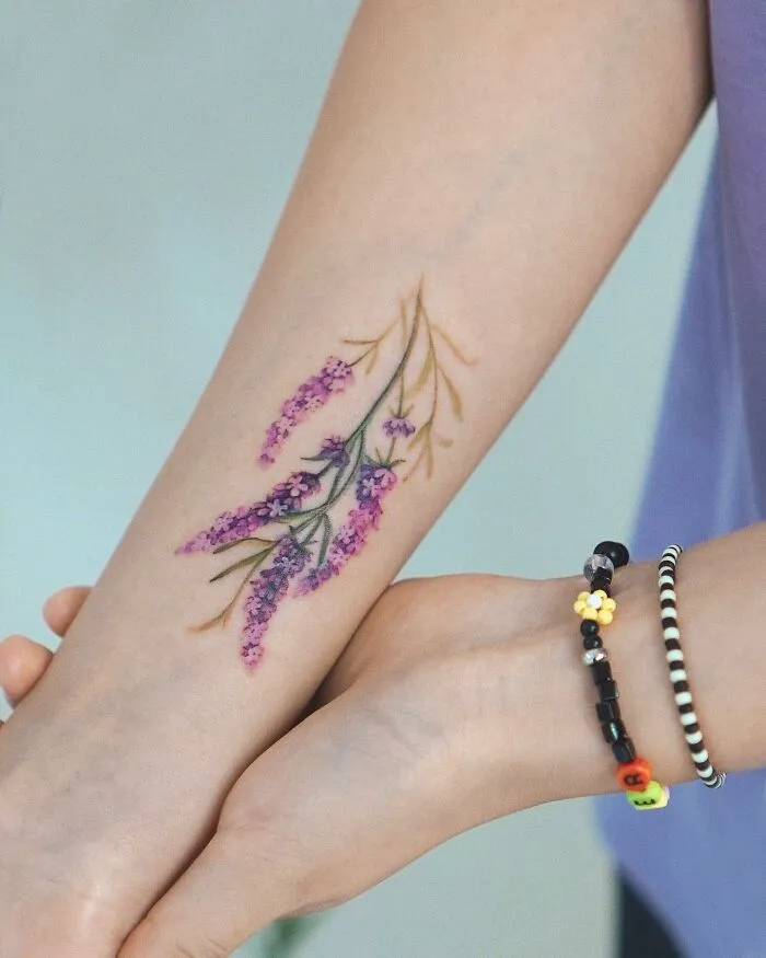 A forearm tattoo with purple flowers, resembling a painting, next to a beaded bracelet.