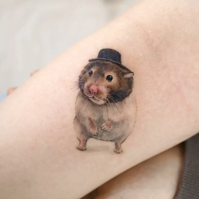 Tattoo of a hamster with a top hat, showcasing painting-like detail on skin.