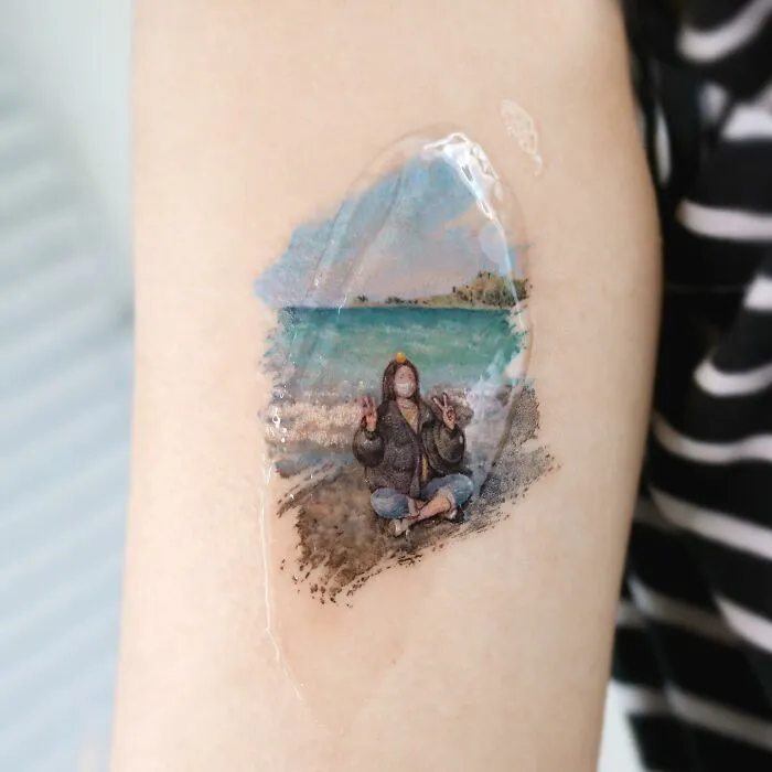 A painting-like tattoo of a person meditating on a beach with a scenic ocean and sky background.