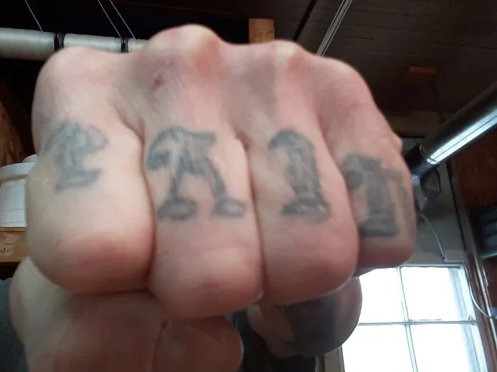 Knuckle tattoos spelling "PAIN" on a clenched fist, shown in a close-up outdoor setting.