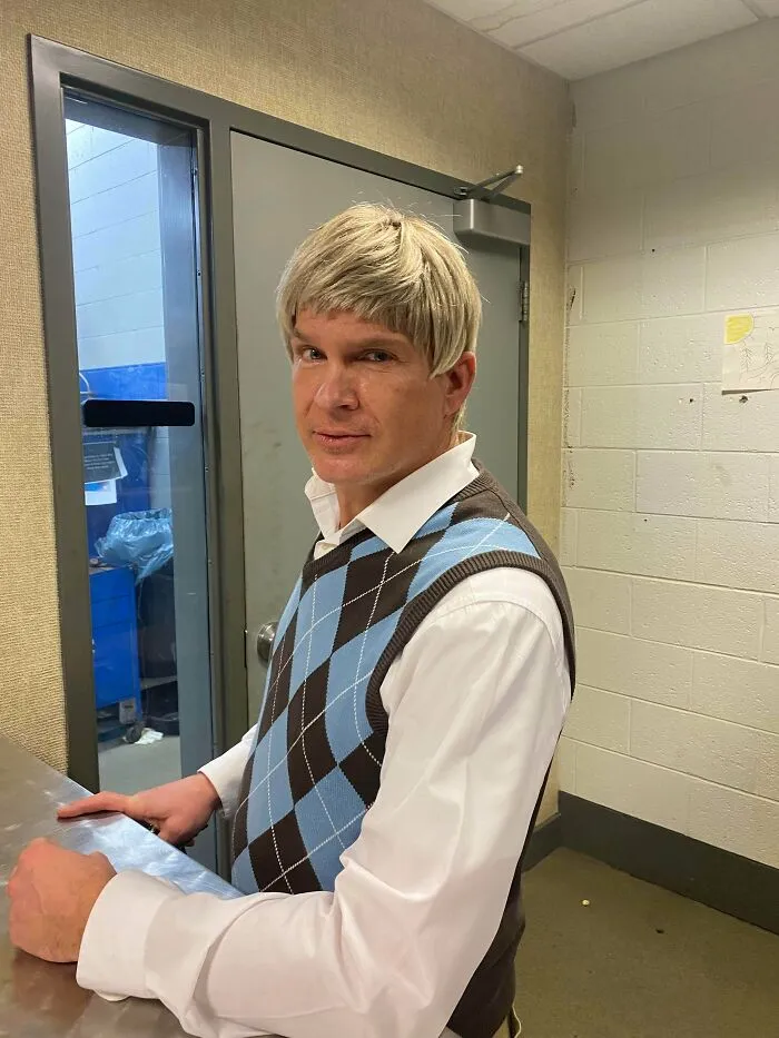 Man with an unusual hairstyle wearing a blue and brown argyle sweater vest, standing indoors. Worst hairstyles showcased.