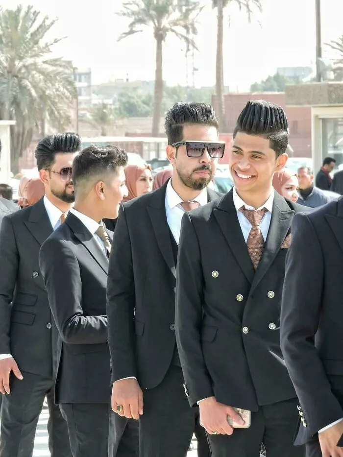 Group of men in suits with unique, styled hairstyles, standing outside and socializing.