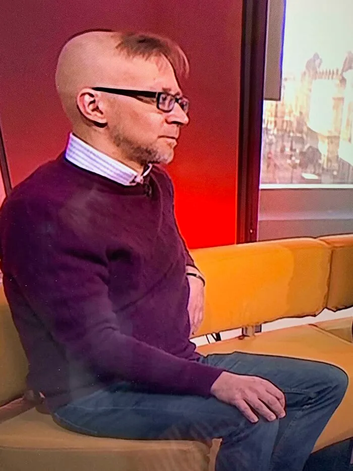 A man with an unusual hairstyle, wearing glasses and a purple sweater, sitting on a yellow couch in a room with red walls.