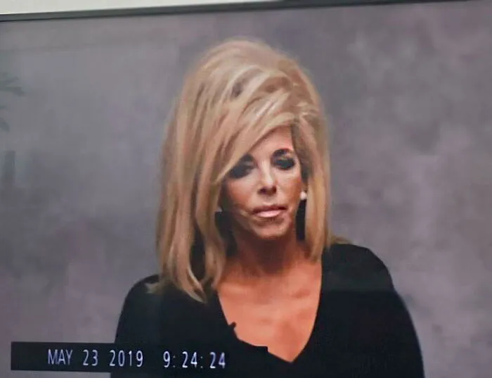 A woman with a voluminous bouffant hairstyle, wearing a black top, in front of a grey background.
