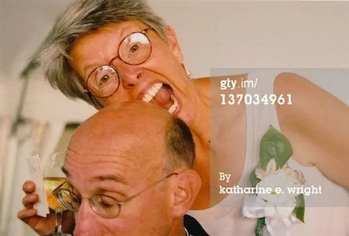 A woman humorously pretending to bite a man's bald head, holding a drink, capturing a weird stock image moment.