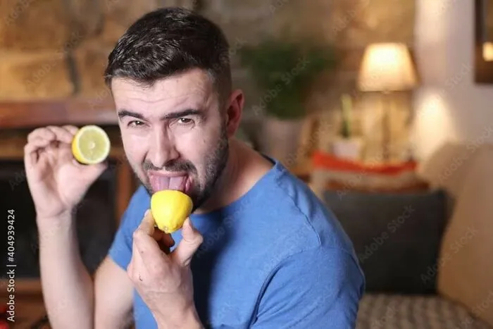 A man in a blue shirt holding lemons, playfully licking one, creating a weird stock image in a cozy room setting.