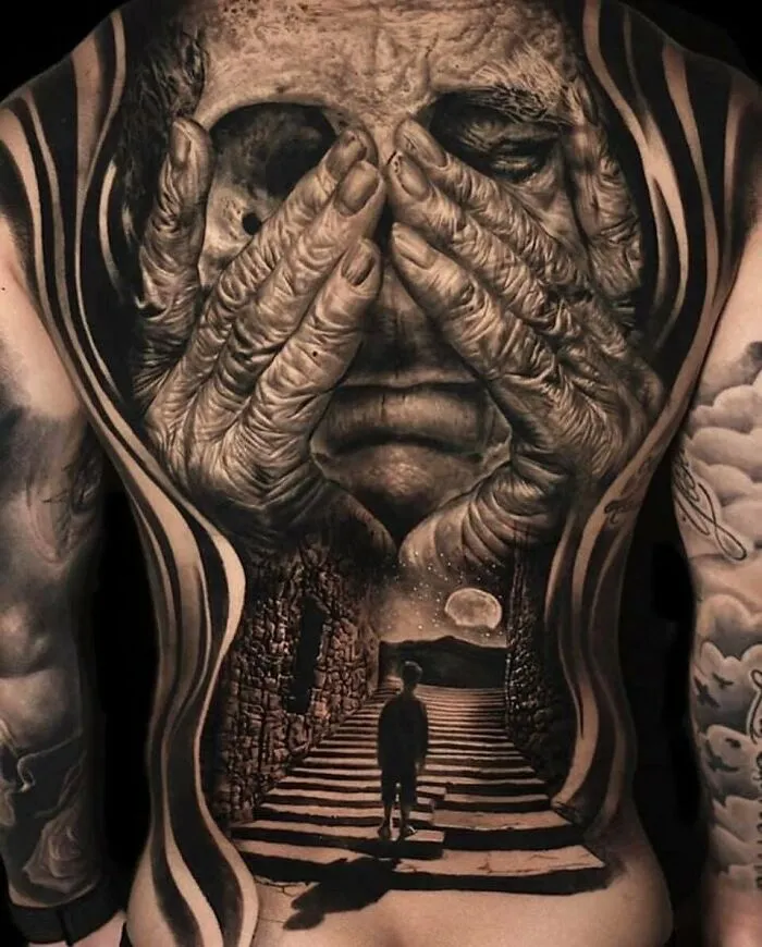 Intricate back tattoo depicting hands covering a face with a surreal landscape scene.