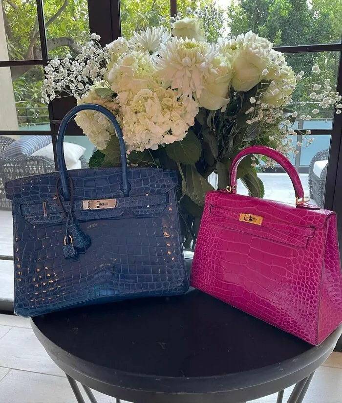 Two luxury designer handbags, one blue and one pink, displayed on a table with a bouquet of white flowers in the background.