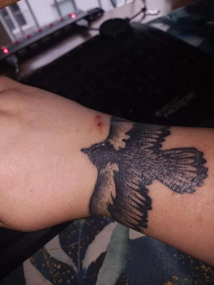 Tattoo of a black bird in flight on a forearm, with a background of a keyboard and window.