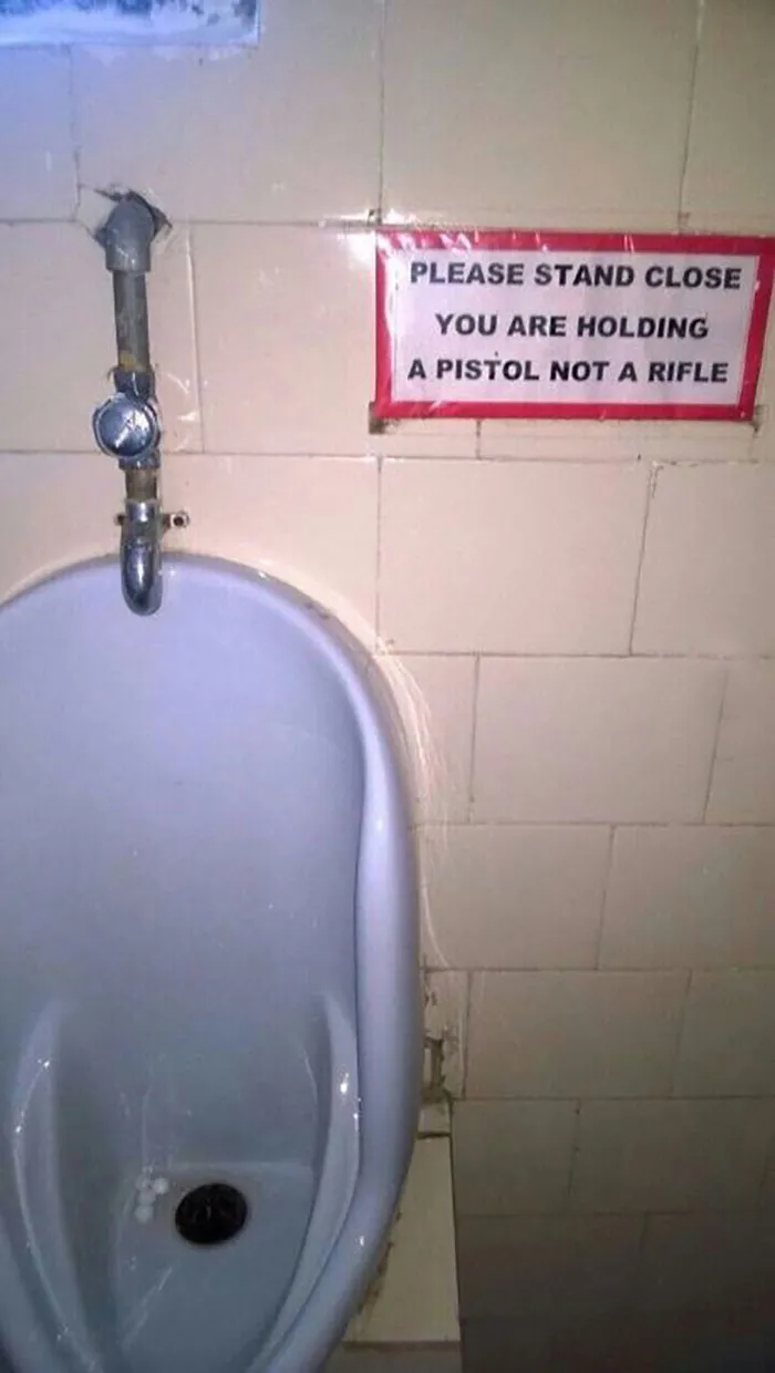 Funny sign above urinal reads, "Please stand close. You are holding a pistol not a rifle," adding humor to the restroom.