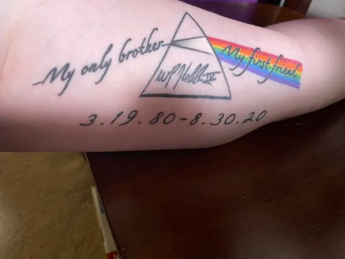 Tattoo on arm featuring a tribute with a triangle, rainbow, and dates, symbolizing a personal memorial.