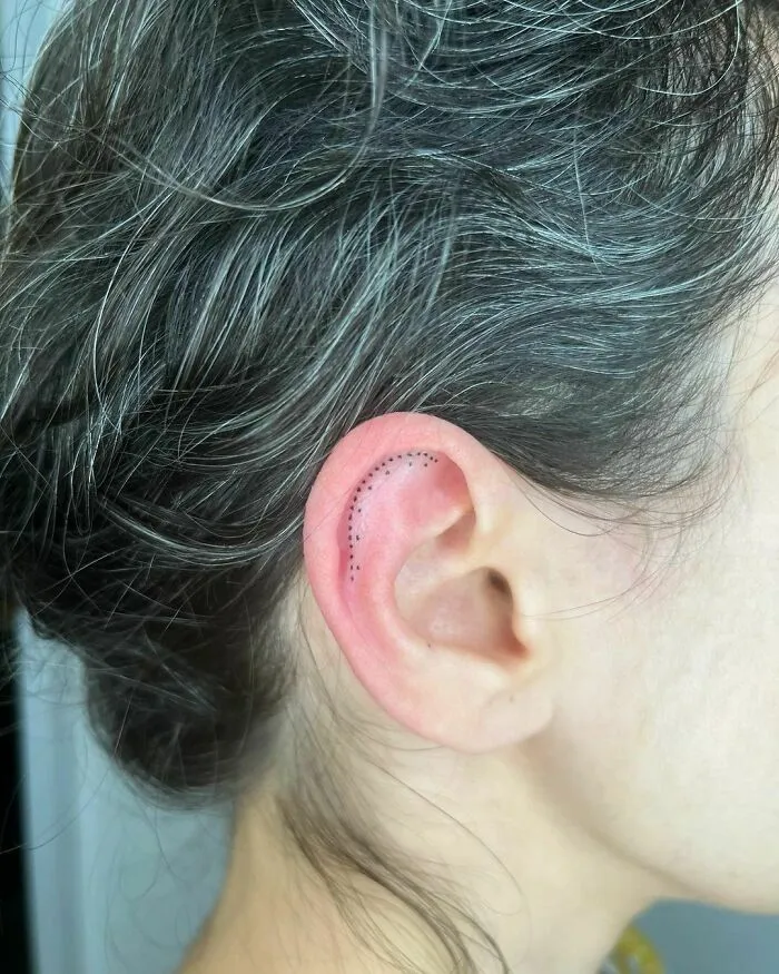 Minimal dot ear tattoo on a woman's inner ear with gray hair.