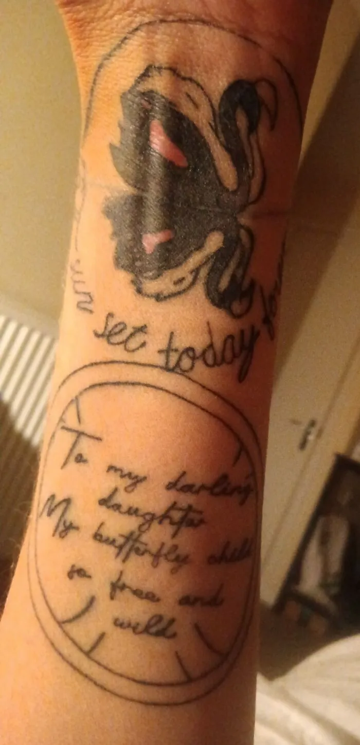 Tattoo of a black and red design on forearm with handwritten script, featuring phrases and a dedication to a daughter.