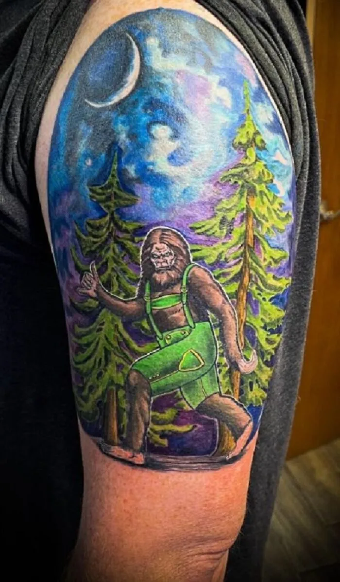 Tattoo featuring Bigfoot in green overalls walking through a forest under a crescent moon.