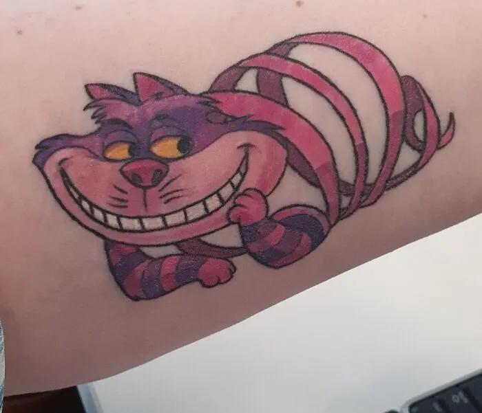 Colorful tattoo of a grinning purple and pink striped cat on an arm.