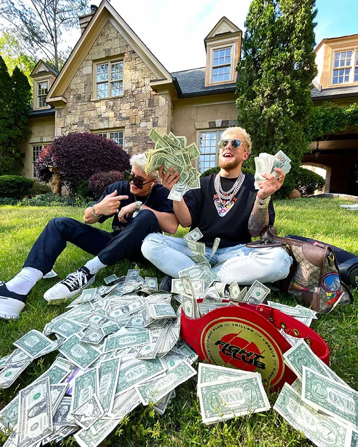 Celebrities flaunting wealth by holding stacks of cash on a lawn, with more money scattered around.