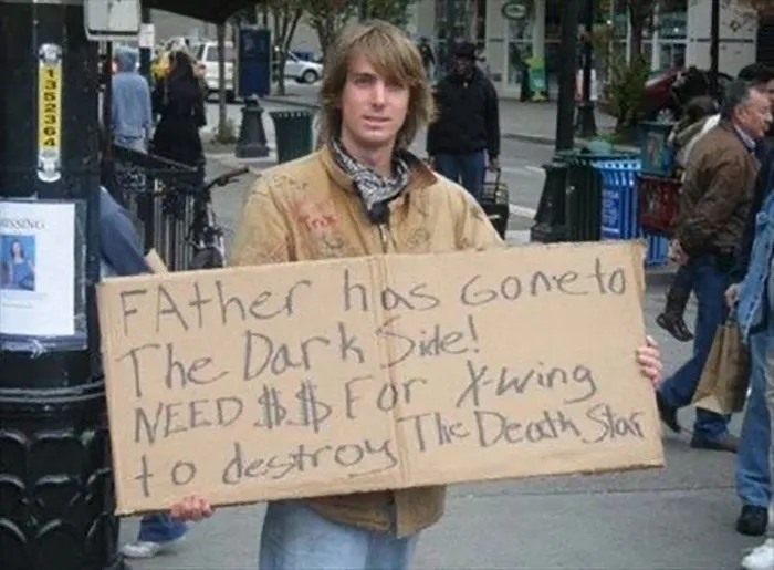 Person holding humorous sign about needing money for X-Wing, referencing sci-fi themes, in a public setting.