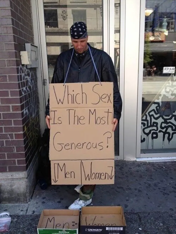 Man holding a humorous sign about generosity with boxes for votes, standing on a city sidewalk.