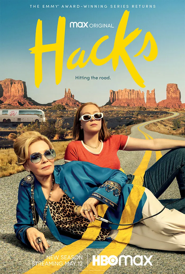 Two women in trendy outfits lounging on a road in a scenic desert, promoting HBO's show "Hacks."