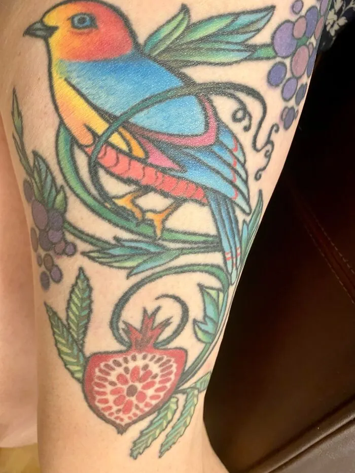 Colorful bird tattoo with floral and fruit designs, featuring a blue and orange bird on skin.