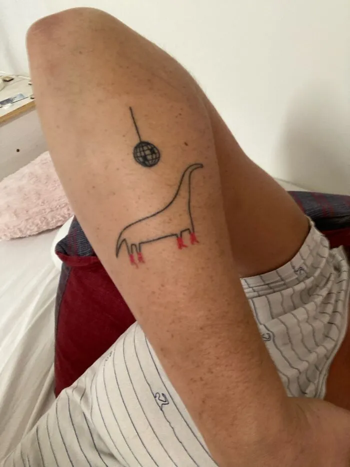 Tattoo of a dinosaur in red boots with a disco ball above, on the back of a person's leg.
