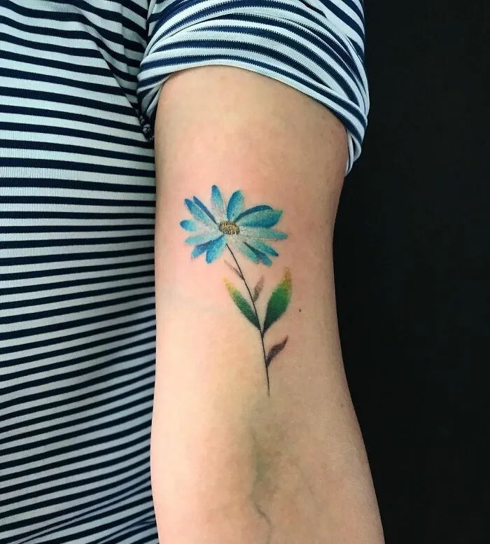 A watercolor tattoo of a blue flower on an arm, showcasing vibrant ink inspiration.