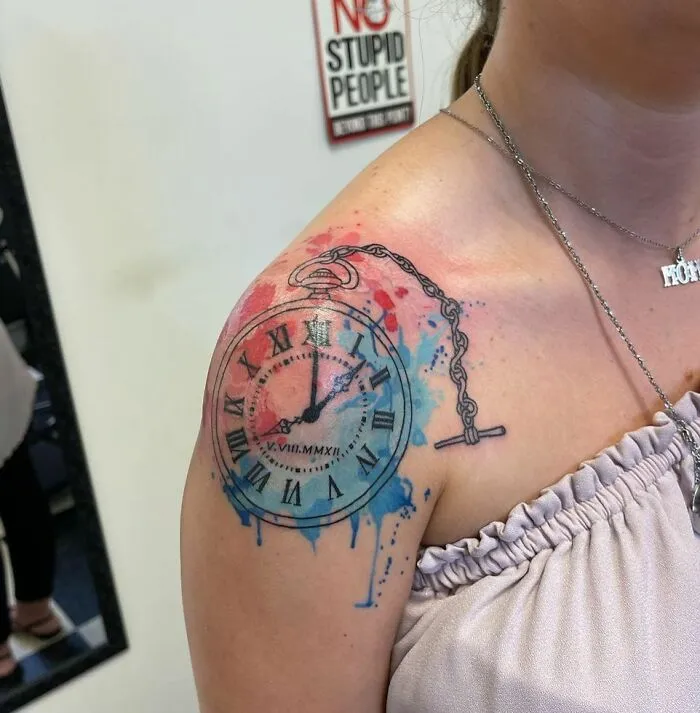 Watercolor tattoo of a clock with vibrant blue and red splashes on a person's shoulder.