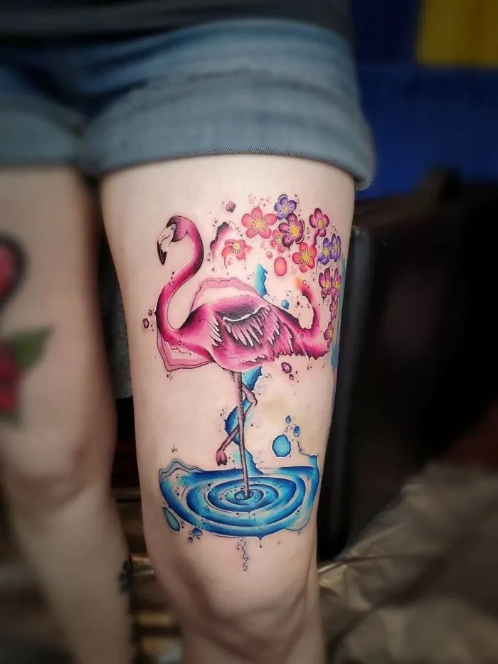 A vibrant watercolor tattoo of a flamingo with floral accents on a person's leg.
