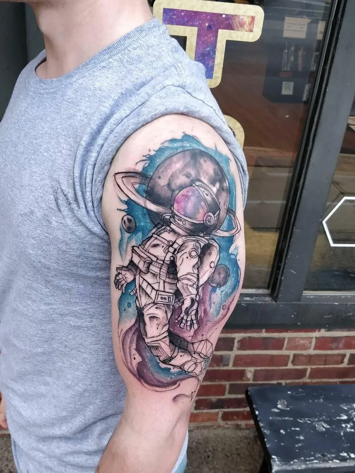 Watercolor tattoo of an astronaut floating in space on a person's upper arm, featuring vibrant colors and cosmic elements.