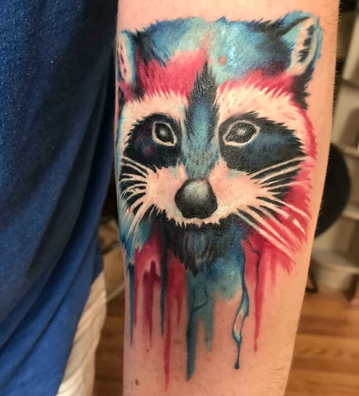 Watercolor tattoo of a raccoon with vibrant blue and red hues dripping down the arm.