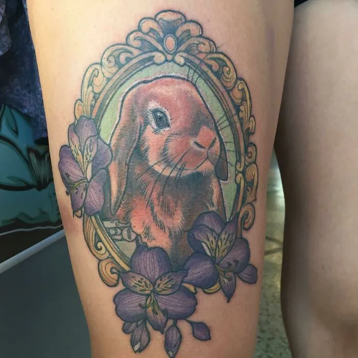Tattoo of a rabbit in an ornate frame with purple flowers, celebrating the pet-human bond.