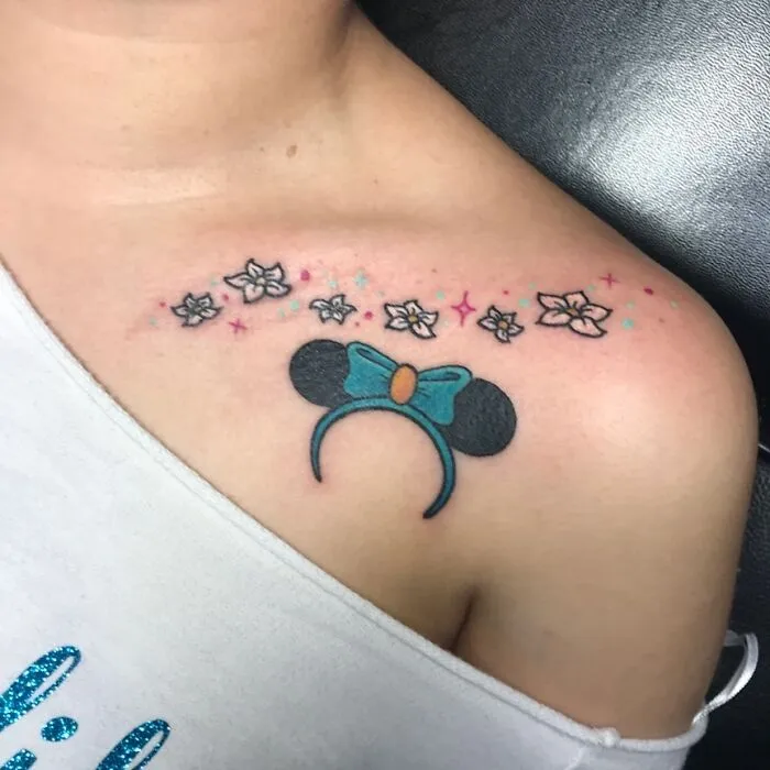 Cartoon tattoo on shoulder featuring a blue bow, mouse ears, and flowers inspired by animated shows.