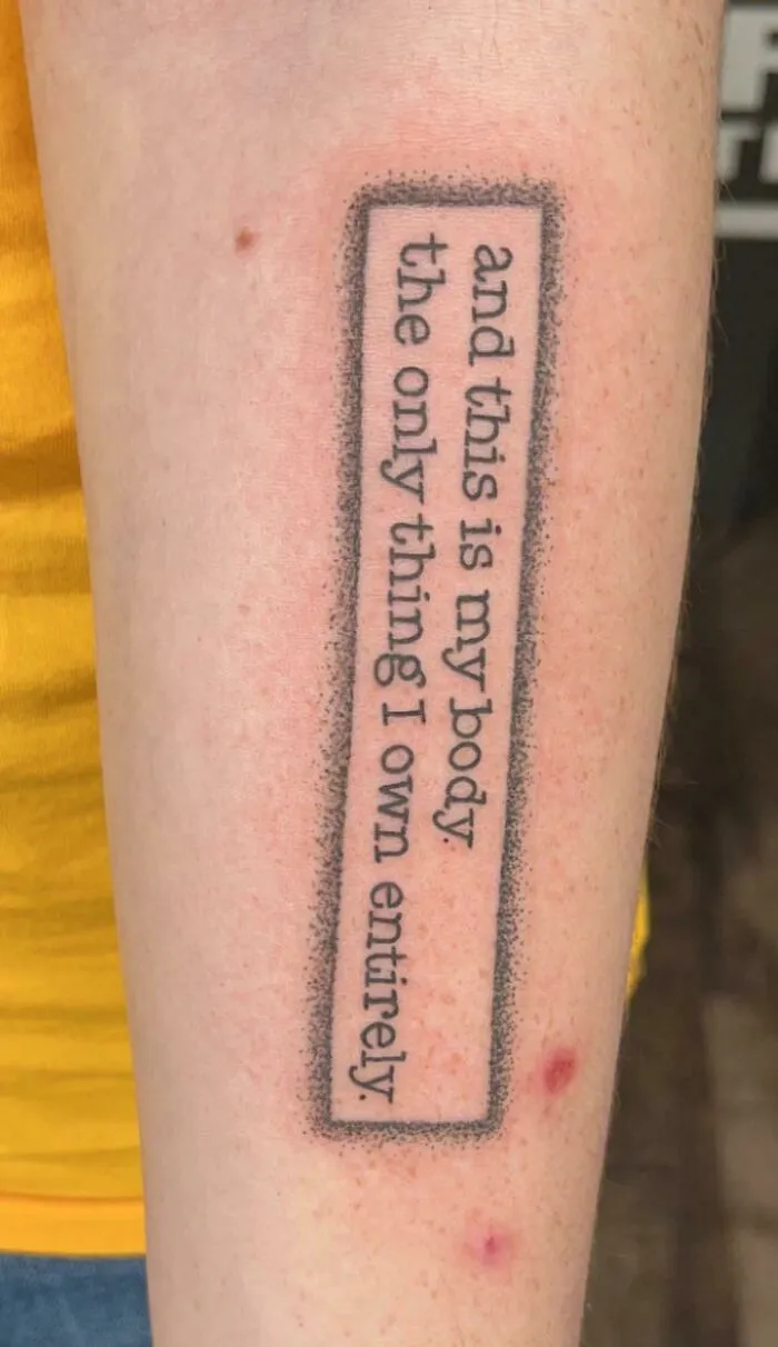 Creative word tattoo on forearm reading "and this is my body the only thing I own entirely," inked in a rectangle frame.