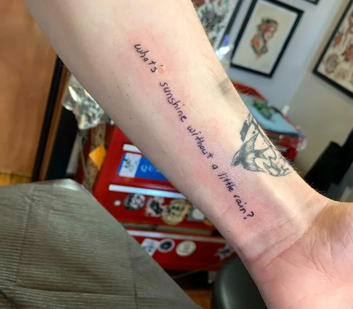 Forearm with a creative word tattoo saying, "what’s sunshine without a little rain?" with surrounding art and decor.
