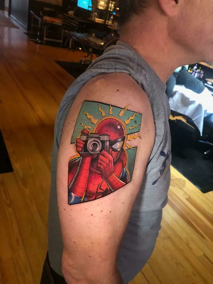 Marvel tattoo of Spider-Man holding a camera on a person's upper arm in a colorful comic style.